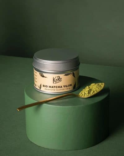 Boîte métal de matcha Yujin bio posée sur un socle vert, cuillère dorée de poudre de matcha